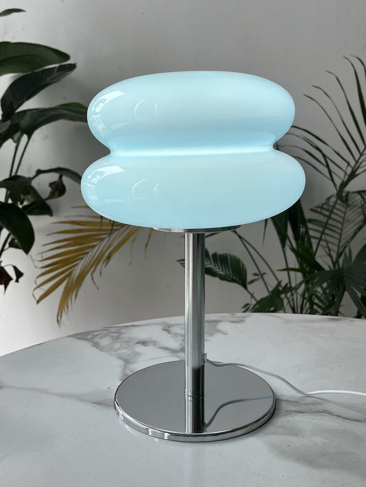 A modern table lamp with a blue, cloud-shaped shade, mounted on a chrome base. The lamp is on a white surface with green plants in the background.