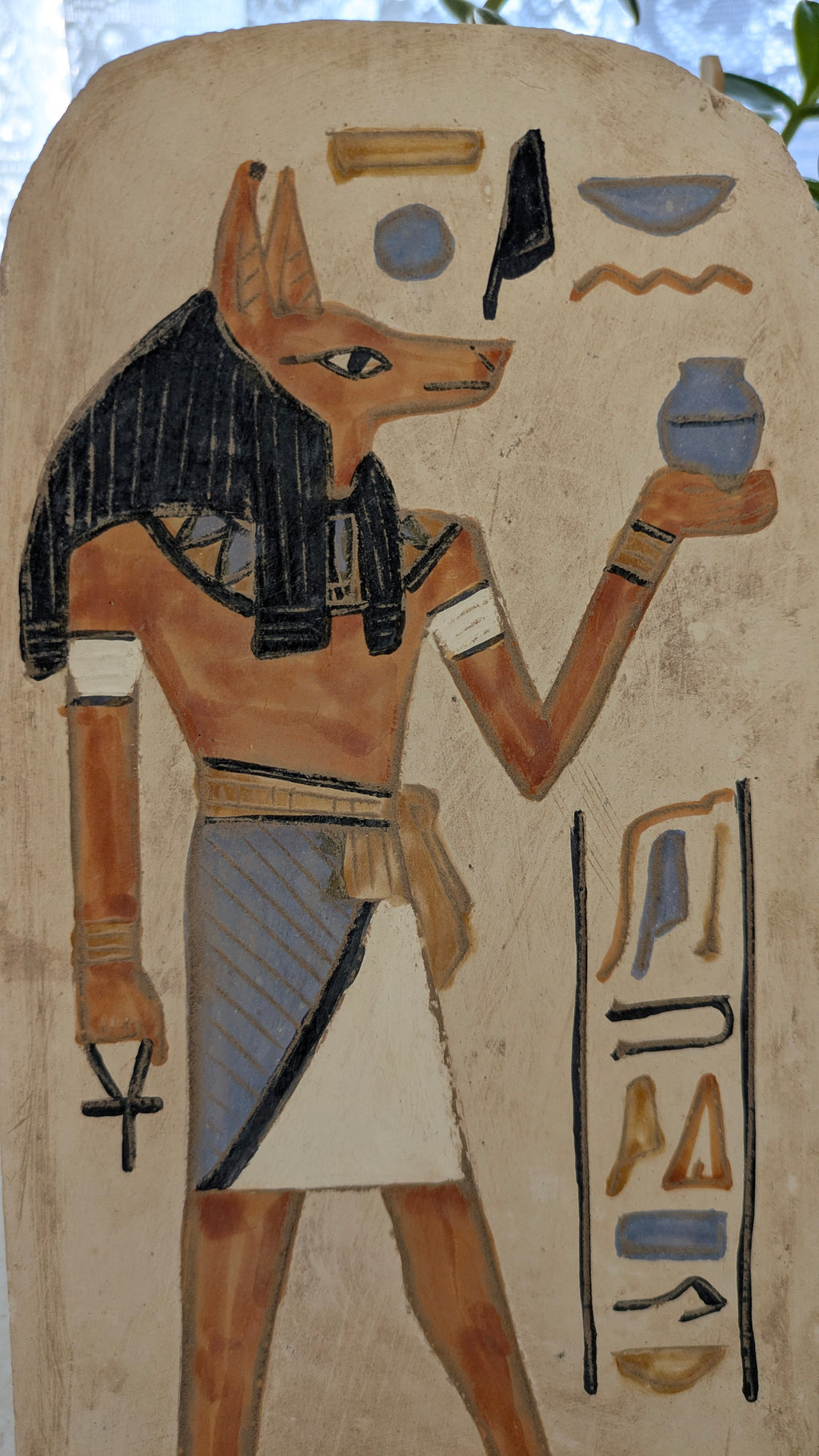 Egyptian deity with a jackal head holding a vase against a plain background