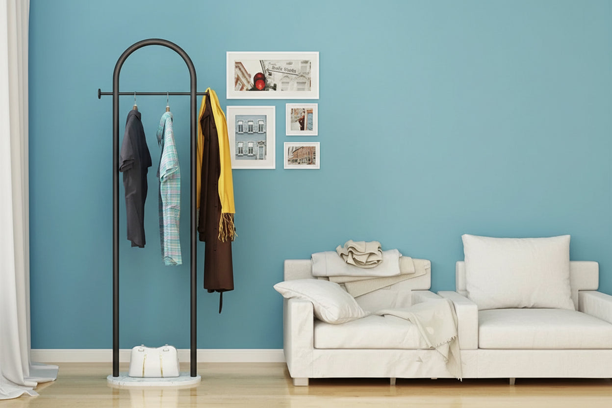Black metal coat rack with clothes against a blue wall with framed pictures, next to a white sofa.
