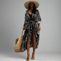 A woman wearing a black and white tie-dye printed beach kimono with long sleeves, paired with a swimsuit and accessories including a wide-brimmed hat and a large straw tote bag.